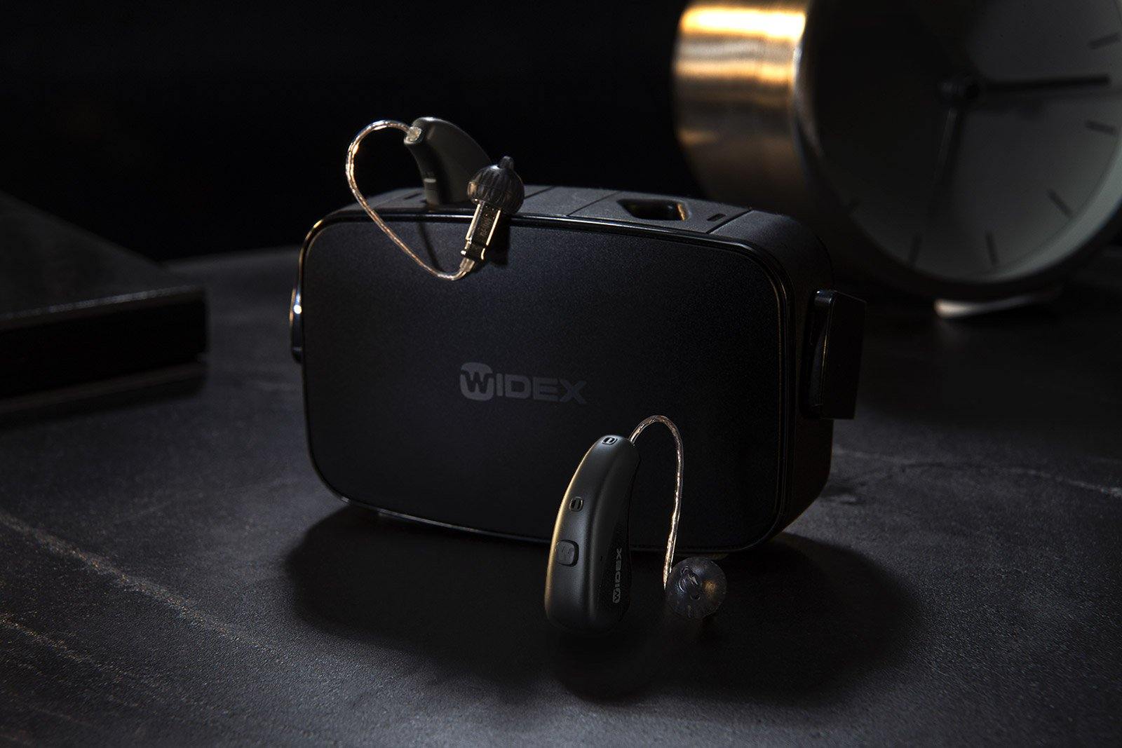 Due apparecchi acustici Widex sono appoggiati davanti a una custodia di ricarica nera su una superficie scura, con un orologio e oggetti sfocati sullo sfondo.