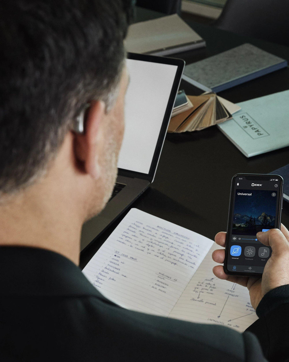 Una persona con un apparecchio acustico utilizza un'applicazione per smartphone mentre prende appunti su un quaderno alla scrivania con un computer portatile e cartelle sullo sfondo.