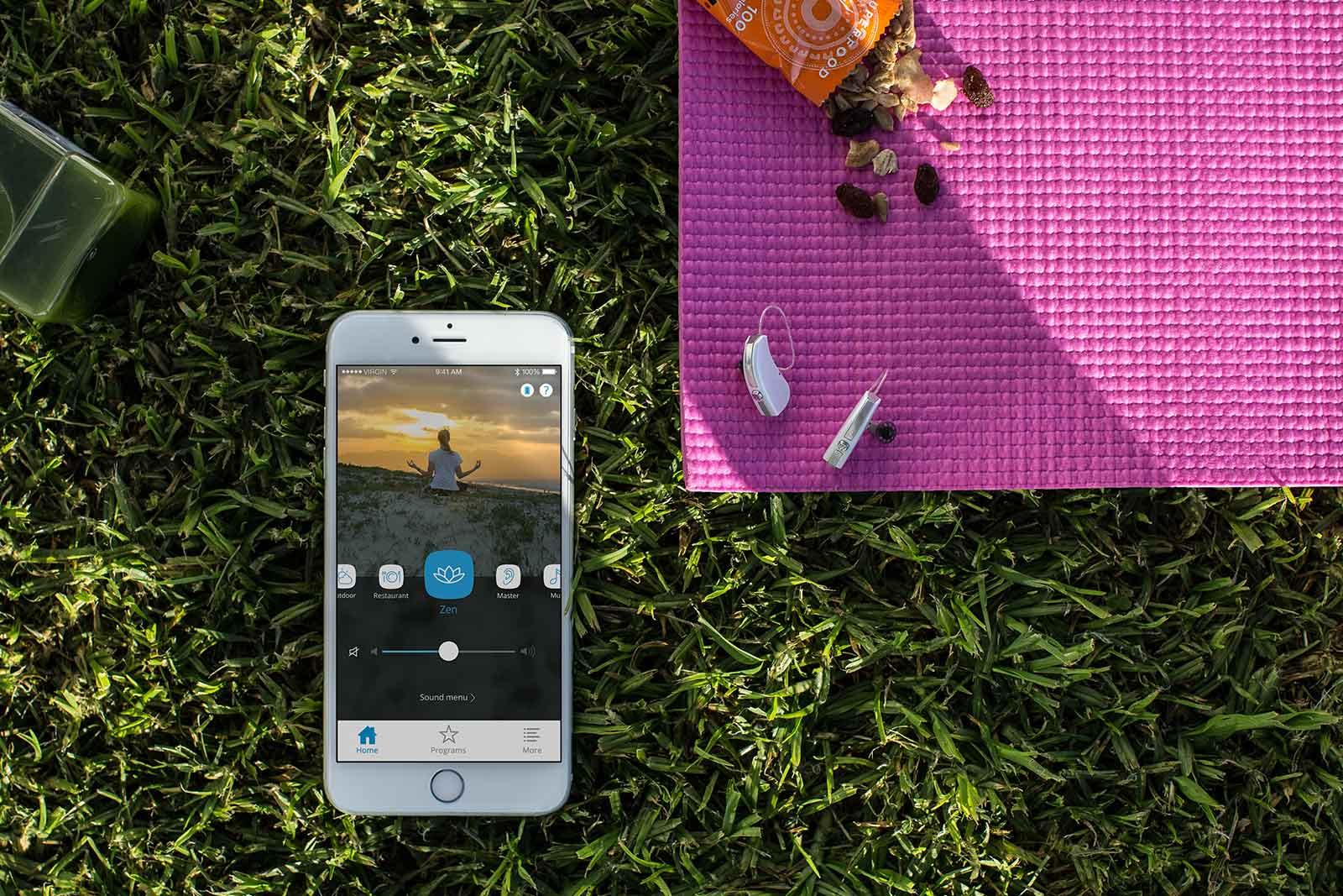 Uno smartphone con un'applicazione per la meditazione giace sull'erba accanto a un tappetino da yoga rosa, un apparecchio acustico, una bottiglia di succo di frutta e una barretta di cereali parzialmente aperta con uvetta e noci.